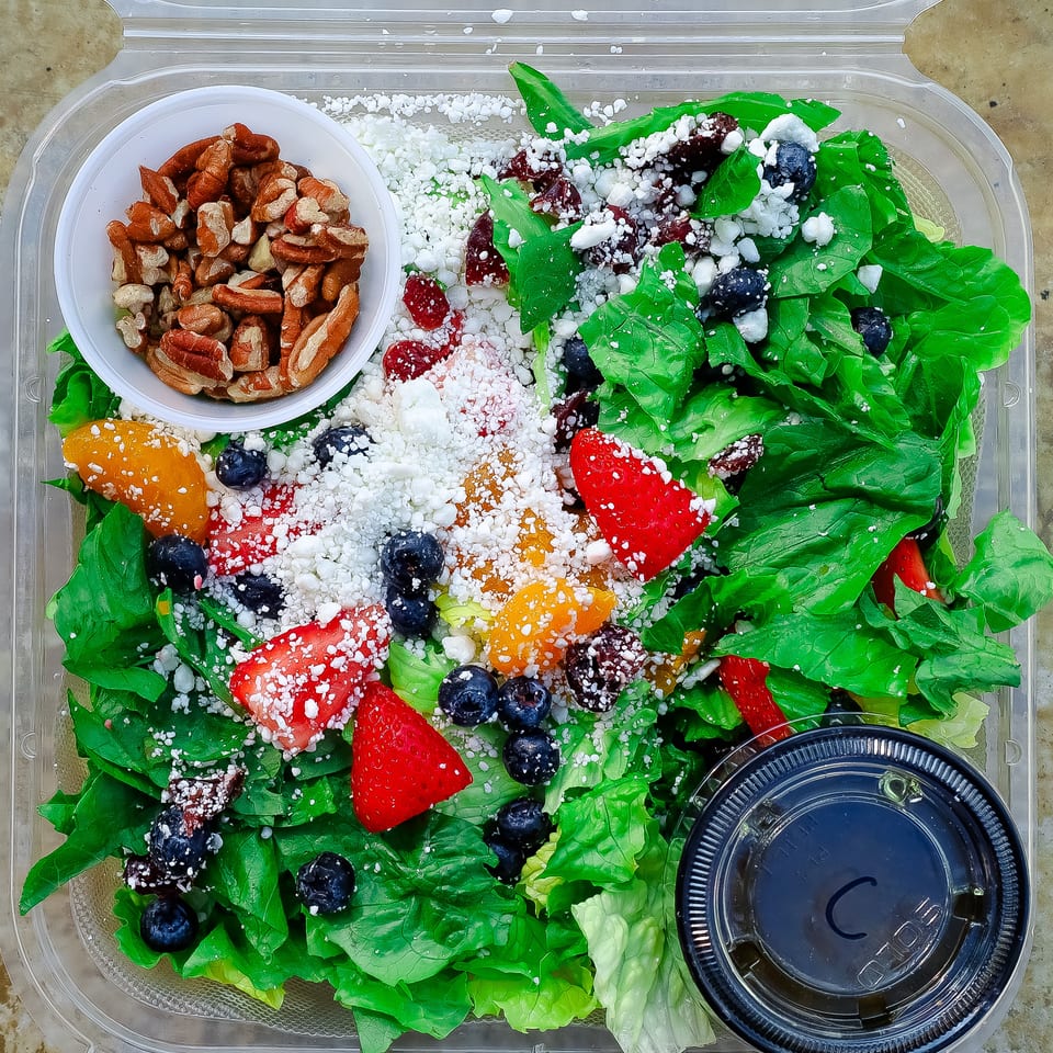 Fresh green salad topped with strawberries, blueberries, mandarin segments, dried cranberries, feta cheese, and pecans in a takeout container.