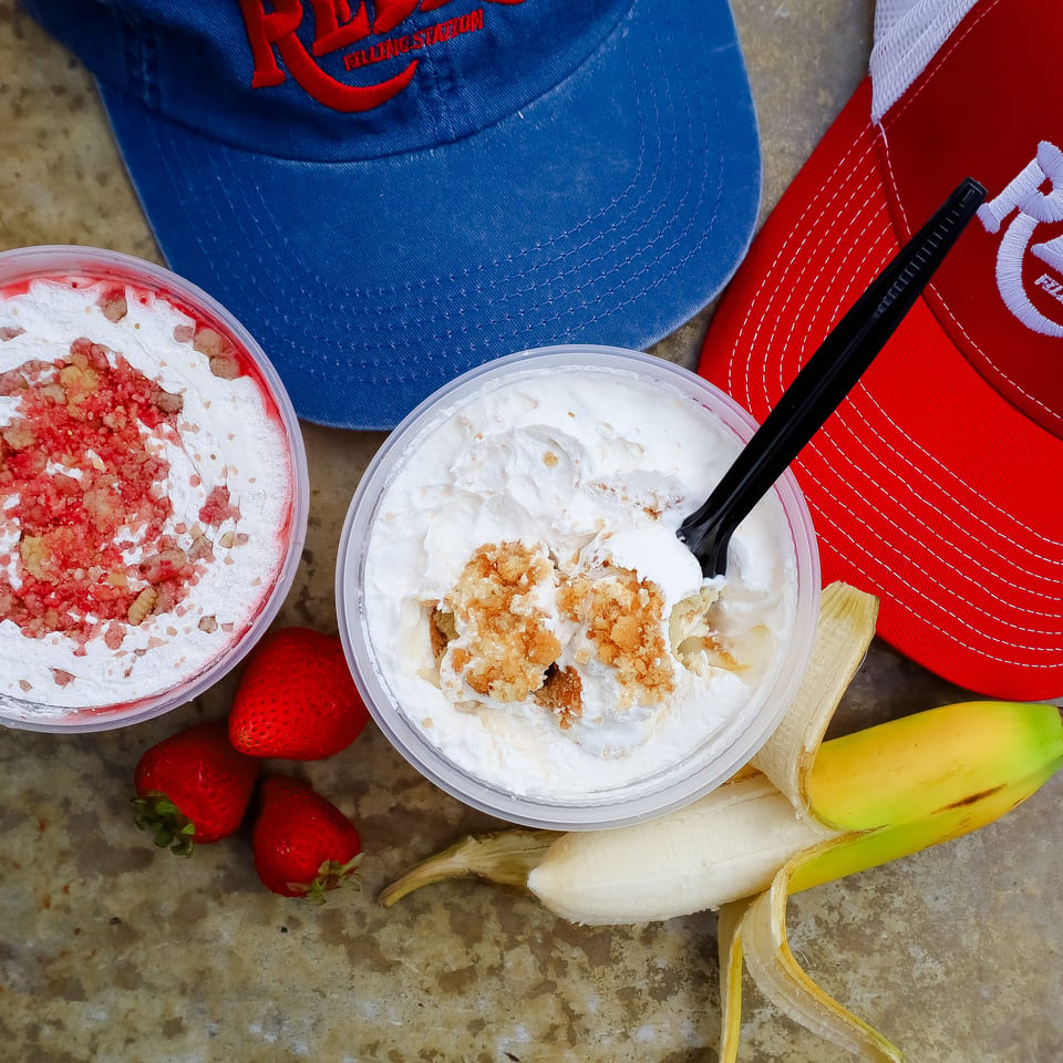 Two whipped-cream dessert cups with crunchy toppings sit beside strawberries, a peeled banana, and colorful branded caps.