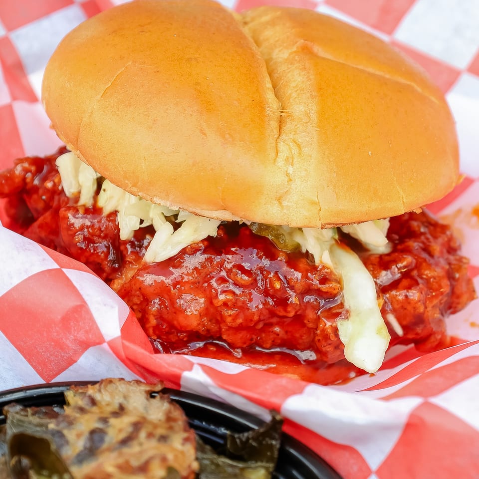 Close-up of a saucy chicken sandwich with lettuce and pickles on a toasted bun, served in red-and-white checkered paper.