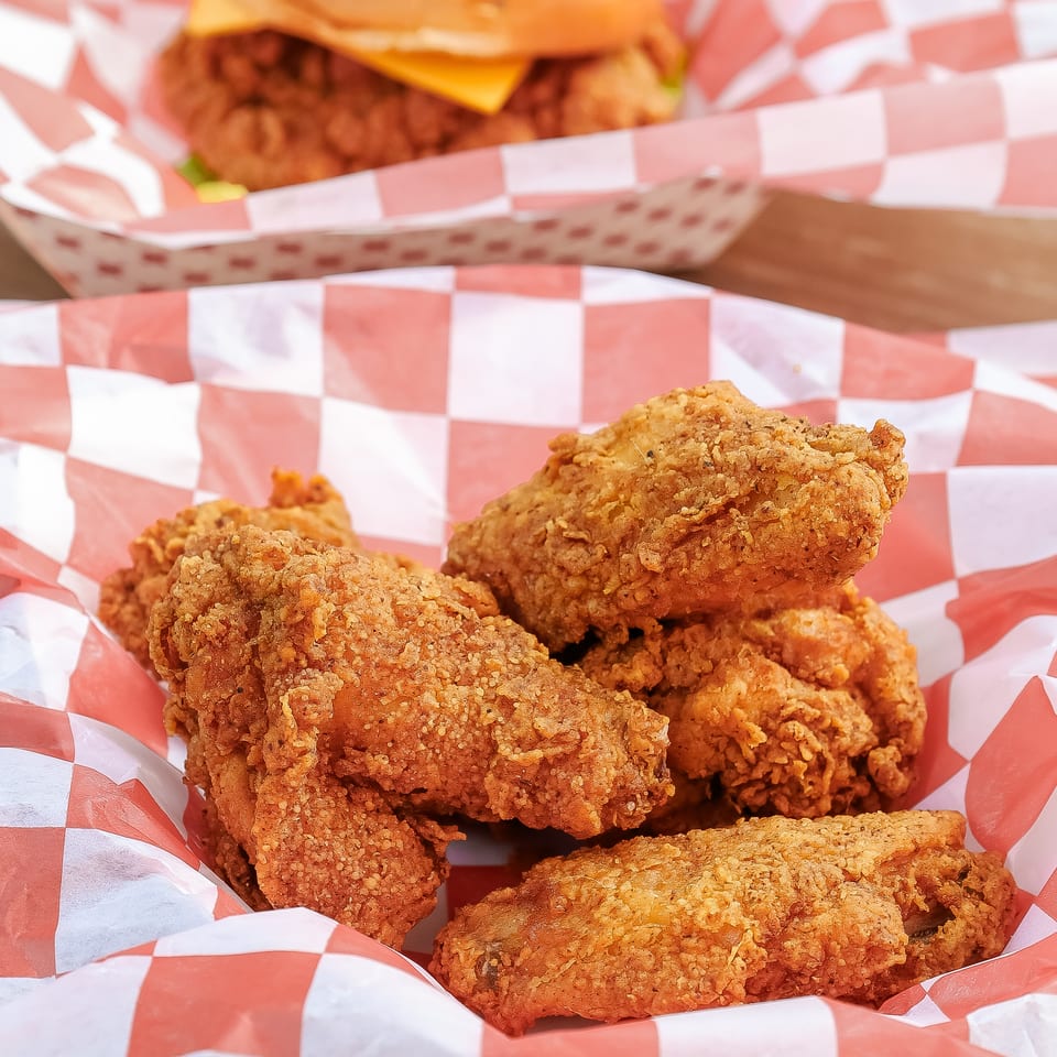 Crispy fried chicken pieces in red-and-white checkered baskets, with a cheeseburger blurred in the background.