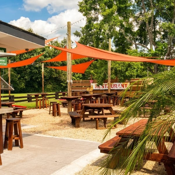 Outdoor restaurant patio with wooden tables, orange shade sails, and a bar area under a blue sky.